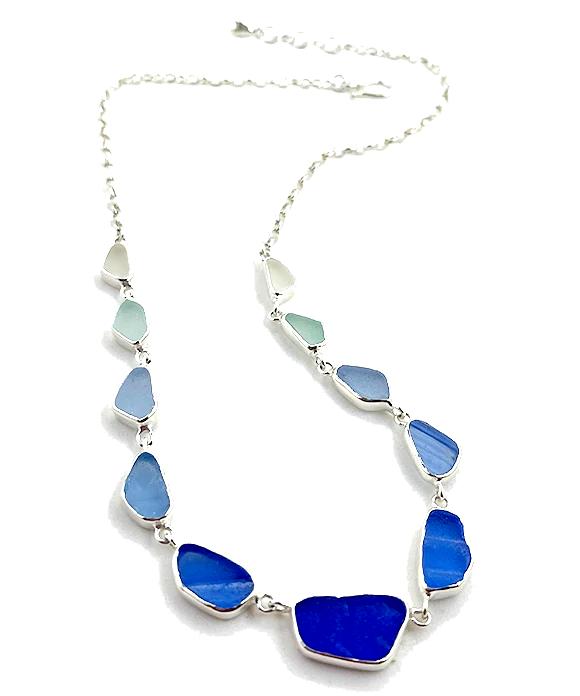 A silver chain necklace featuring a series of irregular, sea-foam glass beads in shades of blue, from pale minty blue to deep royal blue, displayed on a clean white background.