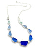 A silver chain necklace featuring a series of irregular, sea-foam glass beads in shades of blue, from pale minty blue to deep royal blue, displayed on a clean white background.